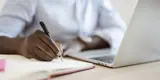 man writing on notepad in front of laptop